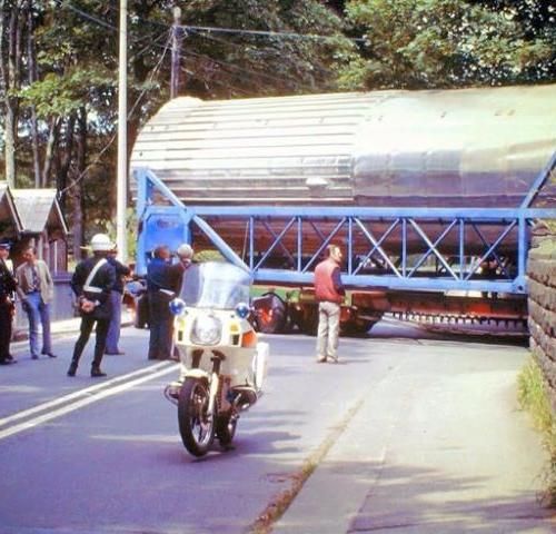 vintage image of a motorbike blocking the road, behind are police and staff supporting transport of a large vehicle into estate